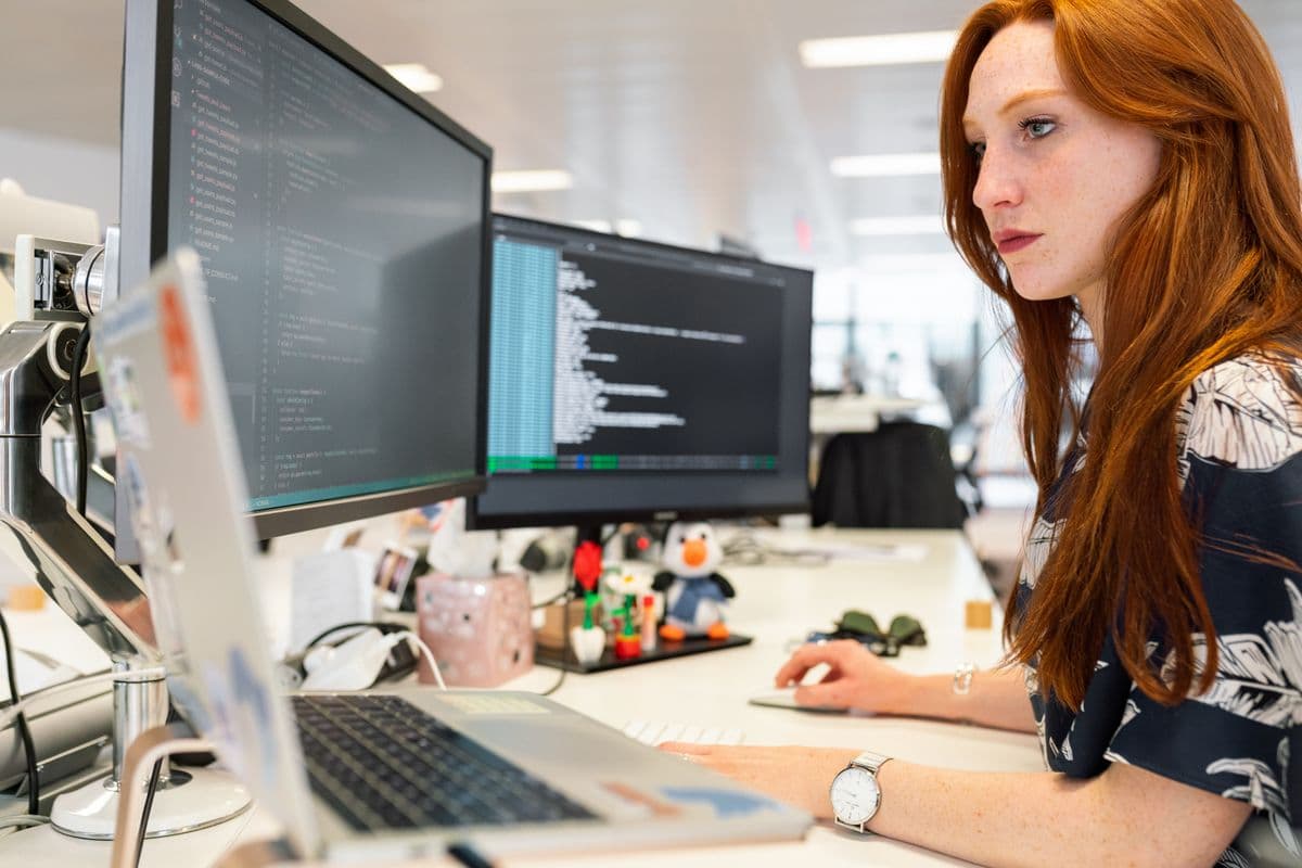 Softwareentwicklerin arbeitet konzentriert an mehreren Bildschirmen mit Quellcode in einem hellen Büro